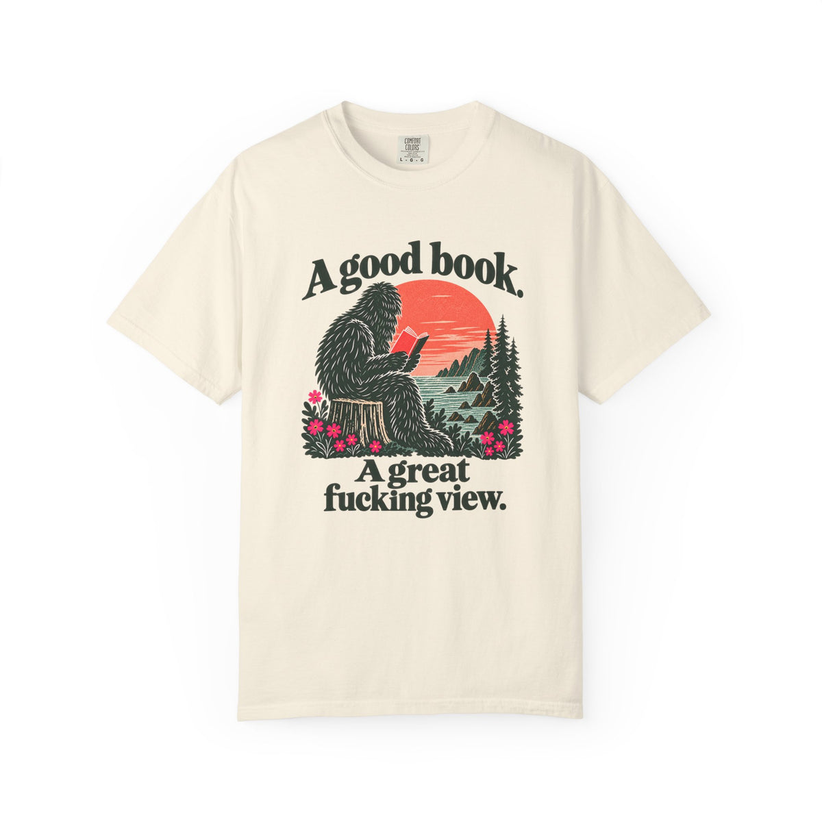 Ivory T-shirt featuring a seated Sasquatch reading beside redwoods and a coastal sunset, with the words A good book. A great fucking view.