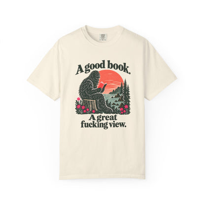 Ivory T-shirt featuring a seated Sasquatch reading beside redwoods and a coastal sunset, with the words A good book. A great fucking view.